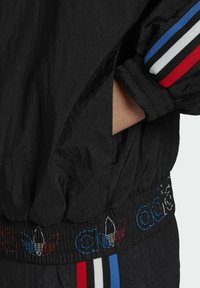 Veste de sport noire avec poignets côtelés ; comporte des rayures rouges, blanches et bleues ainsi que des détails de logo brodés sur la ceinture et la manche.