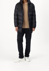 Vadderad svart jacka med huva, beige stickad tröja och mörka randiga byxor. Vita sneakers fulländar outfiten.