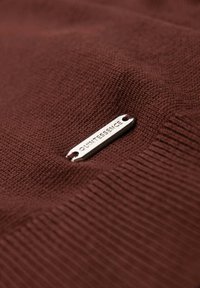 Bruin gebreid stof met een geribbelde textuur, voorzien van een zilveren label met de tekst "QUINTESSENCE" dat is bevestigd door twee kleine gaatjes.