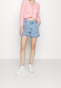 Różowy krótki kardigan z teksturowanym wzorem i złotymi guzikami, w połączeniu z dżinsowymi szortami z wysokim stanem, które mają przednie kieszenie i jasny odcień.