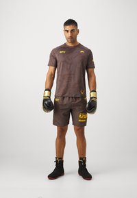 Brahno potiskan nastopni T-majici in kratkih hlačah z logoma UFC in Venum. Črne boksarske rokavice in čevlji z zlatimi detajli.