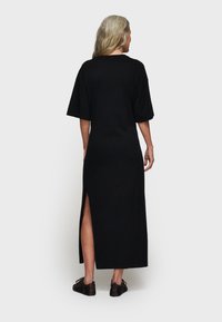 Czarna długa sukienka z krótkimi rękawami, bocznym rozcięciem i luźnym krojem. Gładki materiał o zwiewnym fasonie, noszona z ciemnymi, płaskimi butami.