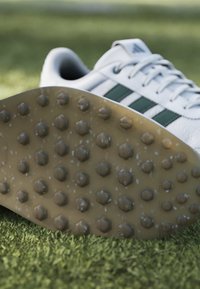 Λευκά παπούτσια Adidas με πράσινες ρίγες που δείχνουν υφή της σόλας με στρογγυλά τρακτερωτά καρφιά σε φόντο πράσινου γρασιδιού.