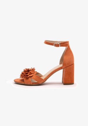 Pomarańczowy zamszowy damski sandał na słupku, z paskiem na kostkę zapinanym na klamrę, odkrytymi palcami oraz dekoracyjnym kwiatowym detalem na przednim pasku.