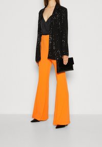 Blazer negro con lentejuelas, top de satén negro, pantalones acampanados naranjas, tacones negros brillantes y un bolso de mano negro con textura.