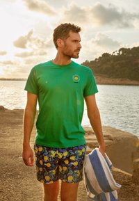 T-shirt en coton vert avec un motif circulaire représentant un citron, associé à un short bleu décoré de motifs de citrons jaunes. Tenant une serviette rayée bleue.