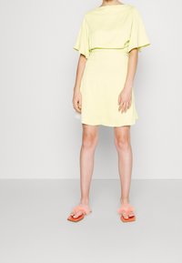 Conjunto de dos piezas amarillo que incluye una blusa corta y holgada de manga corta y una falda a juego. Accesorios con sandalias naranjas y suaves detalles de piel de color rosa.
