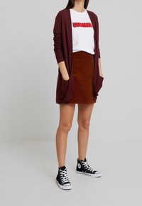 Maroon rippad cardigan med fickor, vit t-shirt med röd text "EQUALITY", brun kordflöjlskjol och svarta höga sneakers.