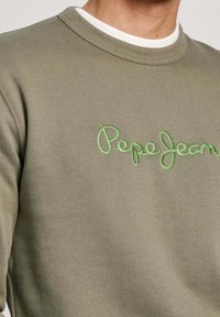 Zbliżenie na khaki bluzę z jasnoszarym haftem "Pepe Jeans" na piersi, noszoną na białej koszulce z okrągłym dekoltem.