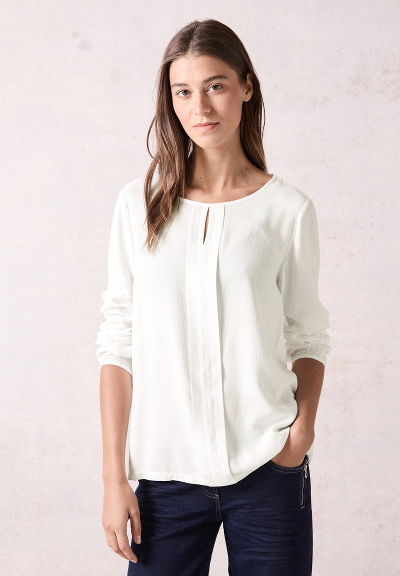 Weiße, strukturierte Langarmbluse mit einem Schlüssel-Loch-Ausschnitt und Faltendetails. Kombiniert mit dunkelblauen Jeans. Einfaches, sauberes Design.