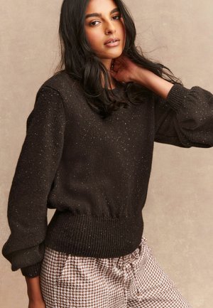 Schwarzer Strickpullover mit gerippten Bündchen und Saum, der ein strukturiertes Muster mit dezenten metallischen Sprenklern aufweist. Kombiniert mit karierter Hose.