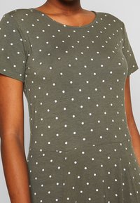 Oliwkowa sukienka na krótki rękaw z okrągłym dekoltem, z białymi kropkami i miękkim, teksturowanym materiałem. Posiada dopasowaną górę i rozkloszowany dół.