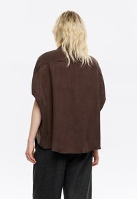 Brown oversized short-sleeve shirt with a rounded hem and smooth texture, paired with gray trousers. Simple, minimalist design.