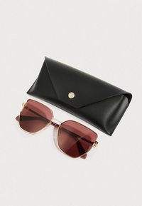 Sončna očala z veliko, kotno kovinsko ogrodje v barvi rožnatega zlata in temno rdečimi lečami. Vključuje črno, trikotno etui z srebrnim zapiranjem na zaskočni gumb.