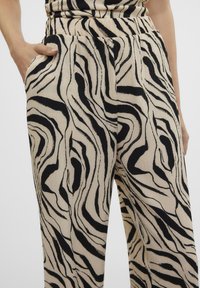 Cremfarbene weite Hose mit einem schwarzen abstrakten Zebra-Muster, aus strukturiertem Stoff und mit seitlichen Taschen. Sichtbares Detail des elastischen Bundes.
