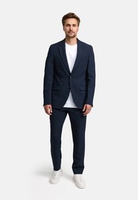 Abito blu navy con giacca a un bottone e pantaloni coordinati, abbinato a una maglietta bianca e sneakers bianche. Tessuto di finitura liscia.