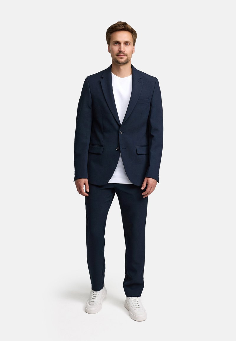 Abito blu navy con giacca a un bottone e pantaloni coordinati, abbinato a una maglietta bianca e sneakers bianche. Tessuto di finitura liscia.