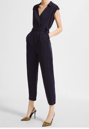 Marineblå jumpsuit med omslagsdesign, korte ærmer og bælte i taljen. Har sidelommer og tilspidsede ben. Bæres med mønstrede højhælede sko.