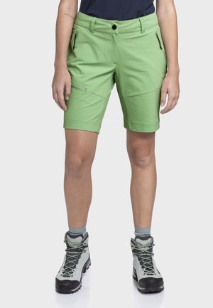 Person trägt hellgrüne knielange Shorts mit Reißverschlusstaschen, hellgraue Wanderschuhe und hellgraue Socken und steht vor einem schlichten Hintergrund.