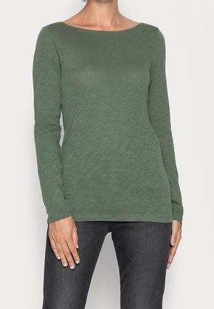 Vrouw draagt een aansluitende olijfgroene top met lange mouwen, gecombineerd met donkergrijze jeans tegen een effen achtergrond.
