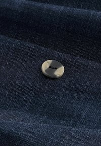 Buton rotund într-un model de broască țestoasă închis, cu două orificii, coase pe o țesătură texturată de culoare închisă. Țesătura prezintă o țesătură fină și o luciu subtil.