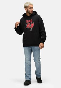Sudadera negra con un logo de los Buccaneers en rojo y blanco. Cuenta con un bolsillo frontal, capucha ajustable, combinada con unos jeans de mezclilla color azul claro y zapatillas negras.