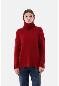 Roter gestrickter Rollkragenpullover mit lockerem Schnitt, gerippten Bündchen und Saum. Die Textur wirkt grob und warm, kombiniert mit dunkelblauen Jeans.