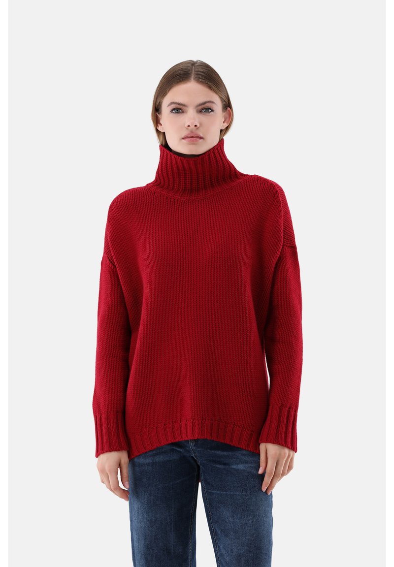 Roter gestrickter Rollkragenpullover mit lockerem Schnitt, gerippten Bündchen und Saum. Die Textur wirkt grob und warm, kombiniert mit dunkelblauen Jeans.