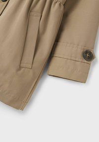 Bež trench kaput s glatkom teksturom, s naglašenim strukom, bočnim džepovima i detaljem na rukavu s gumbima.