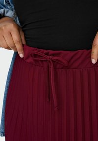 Roke, ki prilagajata zavezano pentljo na pasu nabranega bordo krila, oblečenega z črnim vrhom in jeans jakno.