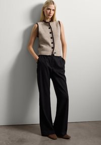 Beige gebreide vest met zwarte afwerking en knopen, gecombineerd met zwarte wijde broek en bruine puntige schoenen, tegen een neutrale achtergrond.