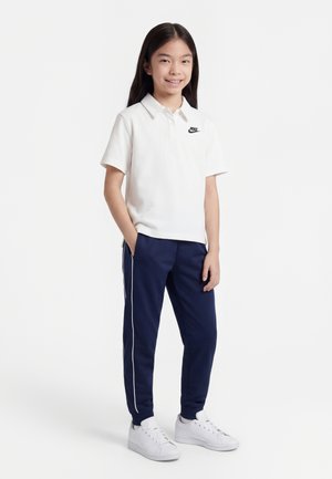 Fille souriante debout, une main dans la poche, portant un polo blanc Nike, un pantalon de survêtement bleu marine avec une bande blanche, et des baskets blanches sur fond blanc.