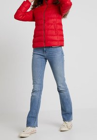 Person i en rød pufferjakke, lyseblå bootcut jeans og hvide sneakers, stående foran en ensartet lys baggrund.