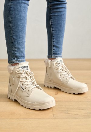 Botas de color crema con cordones y forro de piel, usadas con vaqueros pitillo azules sobre un suelo de madera.