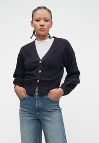 Young person with braided hair wearing a dark navy cardigan with silver buttons over a white top, and light blue jeans with hands in pockets.