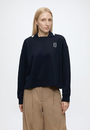 Femeie cu păr blond drept poartă un top bleumarin cu mâneci lungi și guler alb și pantaloni bej largi, stând pe un fundal simplu.