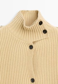 Beige ribbat stickad cardigan med hög krage, två svarta knappar framtill och texturerad tygdetalj.