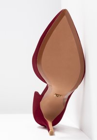 Chaussure à talon haut pointue en suède rouge, inclinée contre un coin blanc, avec une semelle beige et un logo doré près de la voûte plantaire.