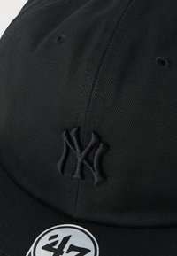 Μαύρο καπέλο μπέιζμπολ με υφασμάτινη υφή, με κεντημένο λογότυπο NY σε μαύρο. Μια κυκλική αυτοκόλλητη ετικέτα με το λογότυπο "47" είναι κολλημένη.