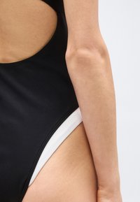 Κοντινό πλάνο ενός ατόμου που φοράει μαύρο μαγιό με λευκό περίγραμμα, δείχνοντας το πλευρικό άνοιγμα καθώς και μέρος του βραχίονα και του κορμού.