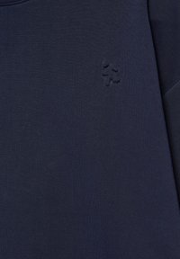 Tissu bleu foncé avec un logo discret en relief constitué de quatre petites formes rondes disposées en forme de croix en haut à droite.