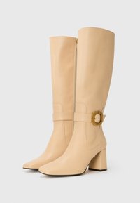 Bottes en cuir beige arrivant au genou avec un orteil carré, un talon carré, une fermeture éclair latérale et une large sangle avec un détail de boucle en ton doré.