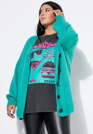 Donna con un cardigan teal, una t-shirt nera con grafiche di occhiali da sole, cocktail, sole e hamburger, e pantaloni in pelle nera.