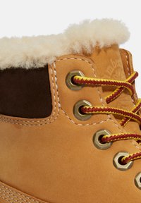 Tanlederstiefel mit einem braunen Wildlederkragen, weichem weißen Fellfutter, Messingösen und roten sowie gelben Schnürsenkeln. Mit weißen Nähten.