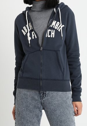 Mujer con sudadera con capucha azul marino con cremallera y cordones blancos sobre un cuello vuelto gris y vaqueros grises claros lavados.