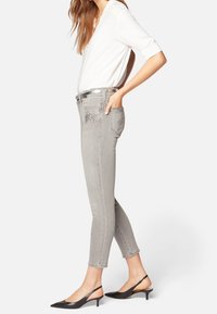 Jeans grises con detalles de tachuelas en los bolsillos delanteros, ajuste de cintura alta y una silueta ajustada y acampanada. Combinados con una blusa blanca y unos zapatos de tacón negro.