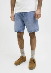 Pantalones cortos de mezclilla, de color azul claro, con corte recto y dobladillo deshilachado. Llevados con calcetines beige y zapatos de ante marrón con cierre de cordones.