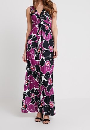 Robe maxi sans manches avec un grand imprimé floral noir, violet et blanc, détail twisté au niveau de la poitrine, portée avec des talons noirs ouverts à l’avant.