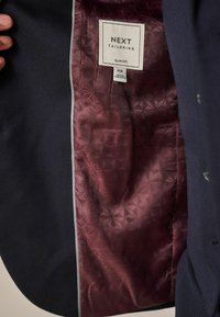 Slim fit marineblauwe blazer met een textuur aan de buitenkant. Gevoerd met een gestreept bordeauxkleurig fabric met een geometrisch ontwerp. Label zichtbaar aan de binnenkant.