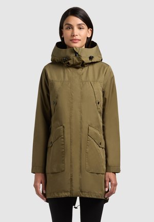 Frau trägt eine olivgrüne Kapuzenparka mit Fronttaschen und Reißverschluss, steht vor einem schlichten hellgrauen Hintergrund.